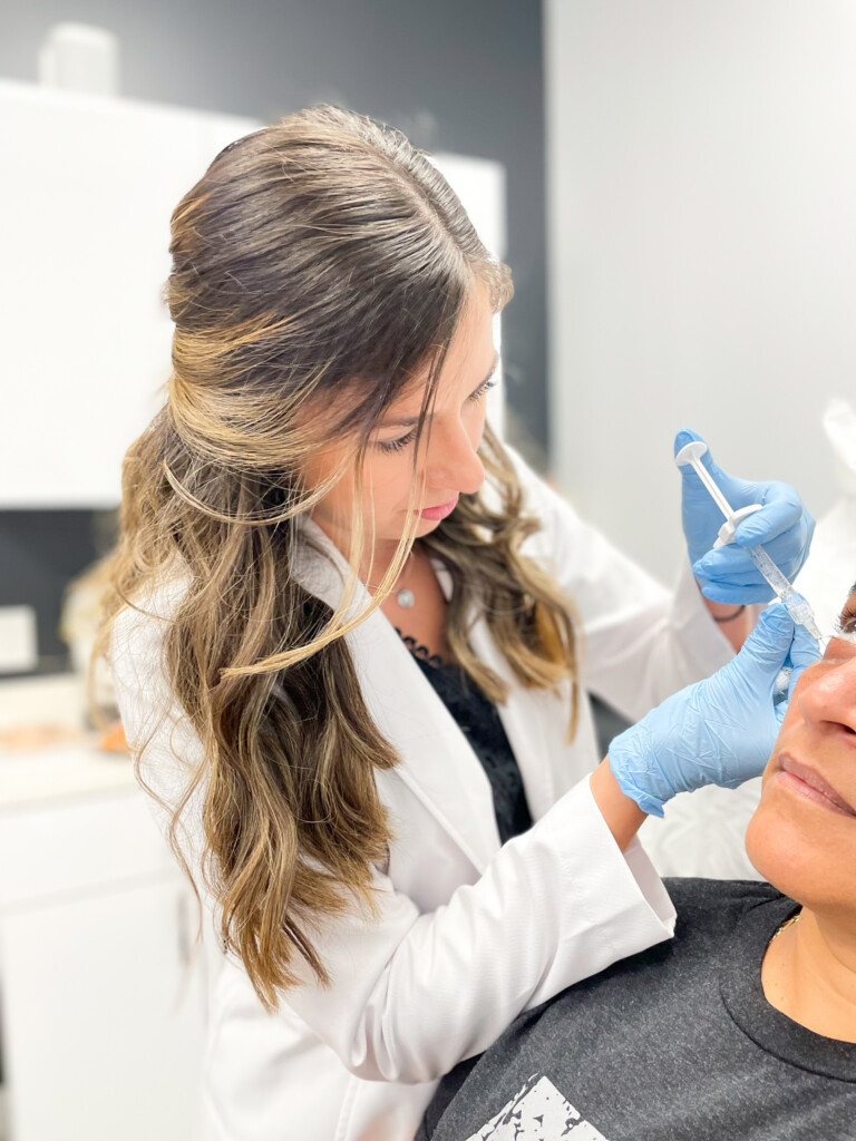 Angela Cusumano, PA-C, wearing blue gloves, performs a facial filler injection near a patient’s under-eye and upper cheek area at Simply Dermatology. Title: Angela Performing Facial Filler Treatment