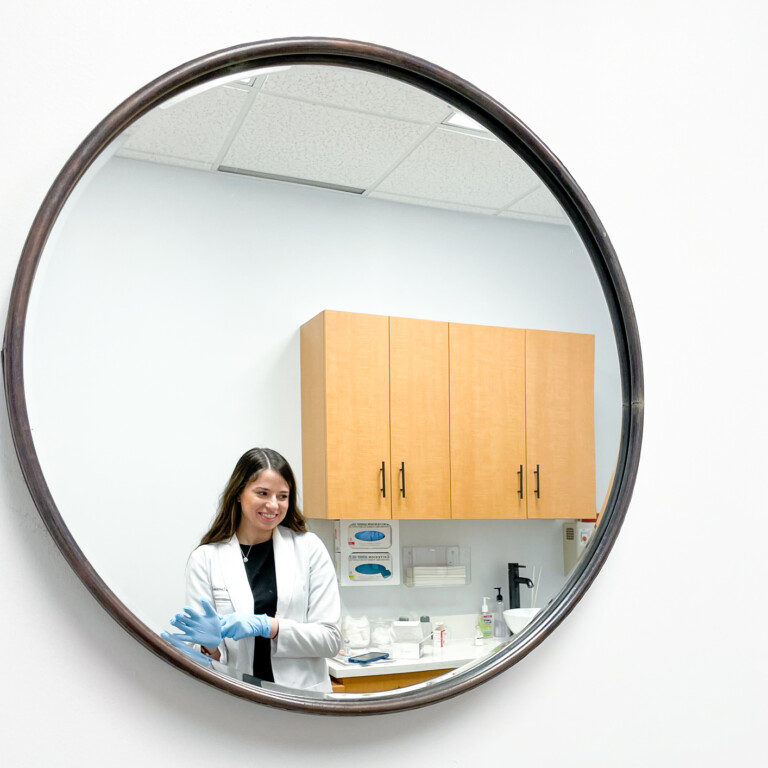 Angela Cusumano, PA, is reflected in a round wall mirror while preparing for a Fraxel Laser treatment in a procedure room at Simply Dermatology, wearing a white coat and blue gloves.