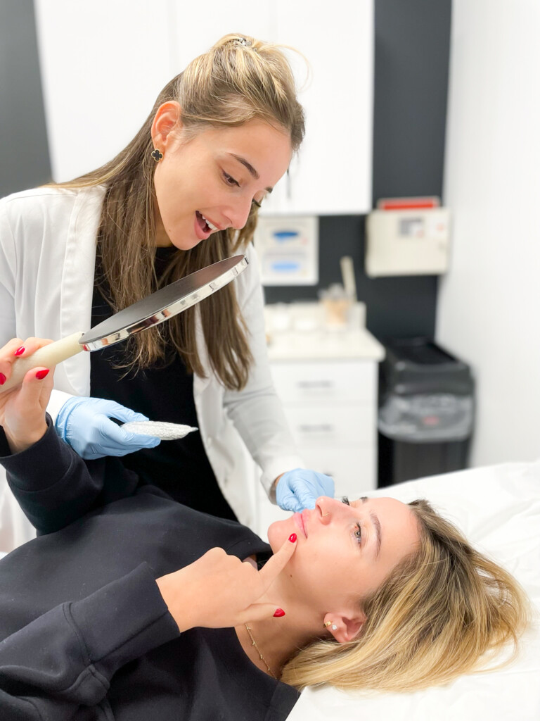 Despina Psyllis, MS, PA-C, holds a handheld mirror while consulting with a patient who points to her chin and lower face before filler treatment.
