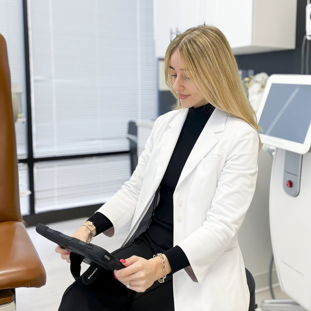 Despina Psyllis, MS, PA-C, seated in a treatment room wearing a white coat and holding a tablet, with an UltraShape Power device beside her.