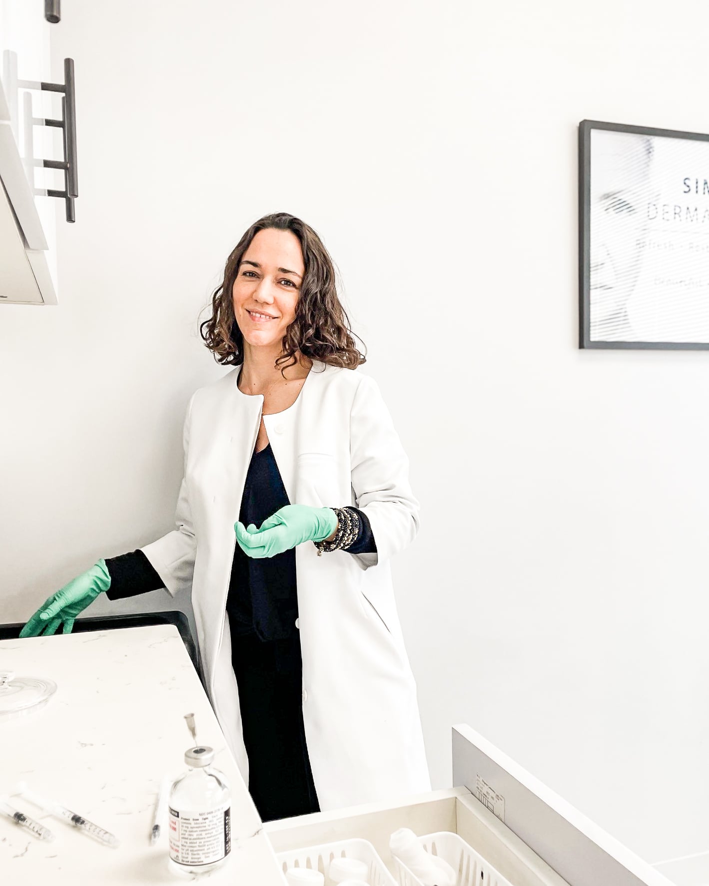 Dr. Kally Papantoniou stands in a Simply Dermatology treatment room wearing a white coat and green gloves beside injectable treatment supplies and syringes.