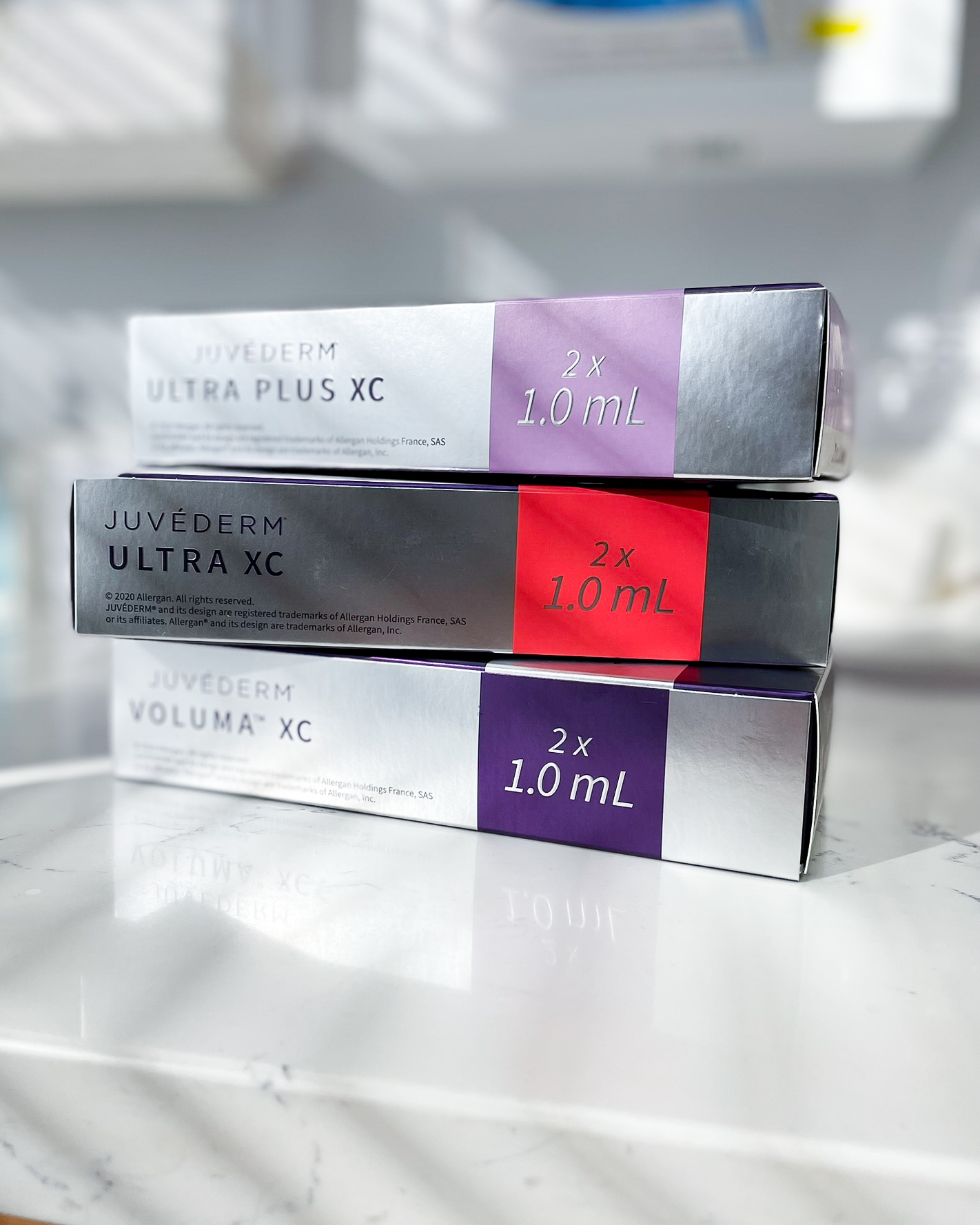 Three stacked Juvéderm filler boxes on a white marble counter in a treatment room. The packages are labeled Juvéderm Ultra Plus XC, Juvéderm Ultra XC, and Juvéderm Voluma XC, each containing 2 x 1.0 mL.