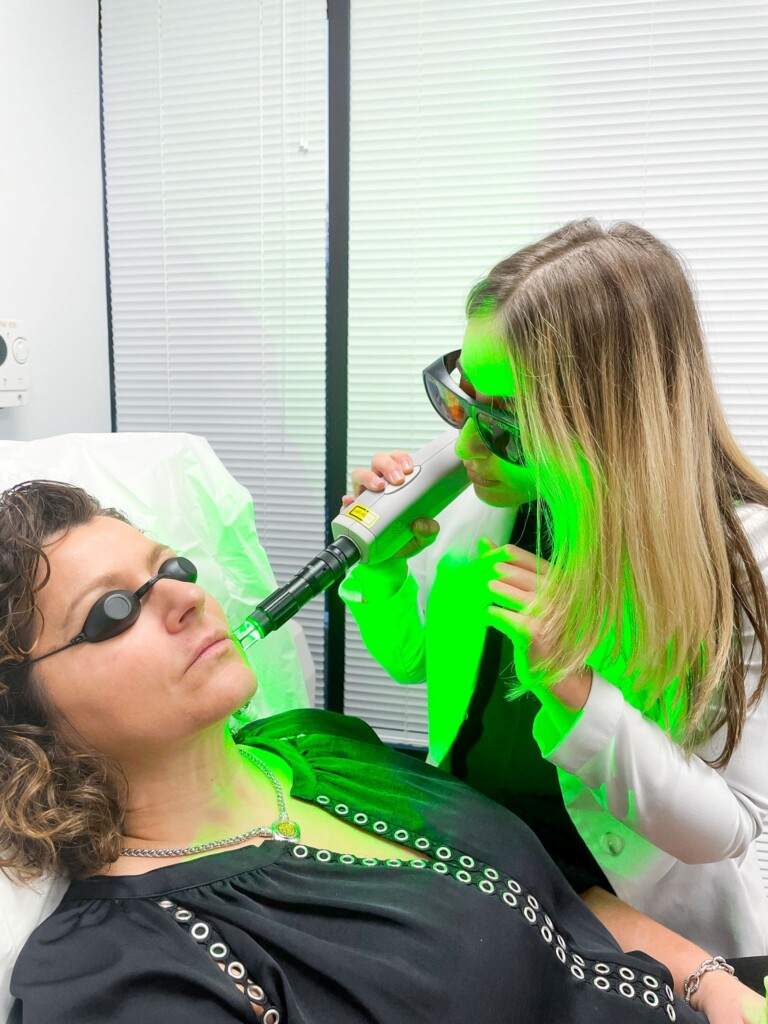 Despina performs a Q-Switch laser treatment on a patient’s face while both wear protective eyewear. A bright green laser light illuminates the treatment area and exam room.