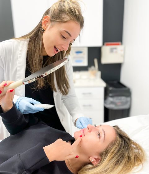 Despina Psyllis, MS, PA-C, holds a handheld mirror while consulting with a patient who points to her lower face before an injectable treatment at Simply Dermatology.