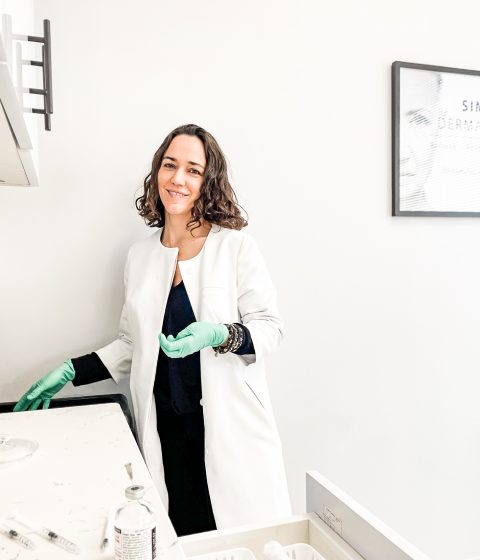 Dr. Kally Papantoniou stands in a Simply Dermatology treatment room wearing a white coat and green gloves beside injectable treatment supplies and syringes.