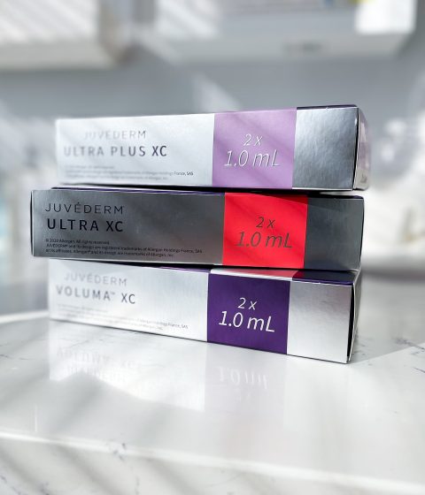 Three stacked Juvéderm filler boxes on a white marble counter in a treatment room. The packages are labeled Juvéderm Ultra Plus XC, Juvéderm Ultra XC, and Juvéderm Voluma XC, each containing 2 x 1.0 mL.