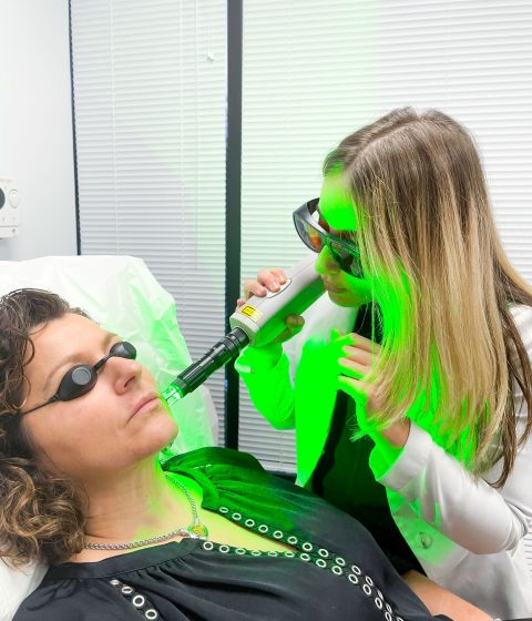 Despina performs a Q-Switch laser treatment on a patient’s face while both wear protective eyewear. A bright green laser light illuminates the treatment area and exam room.